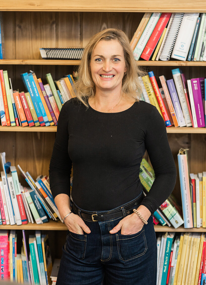 Daniela Burns, Coach in Salzburg, vor einer Bücher-Wand und lächelt in die Kamera.