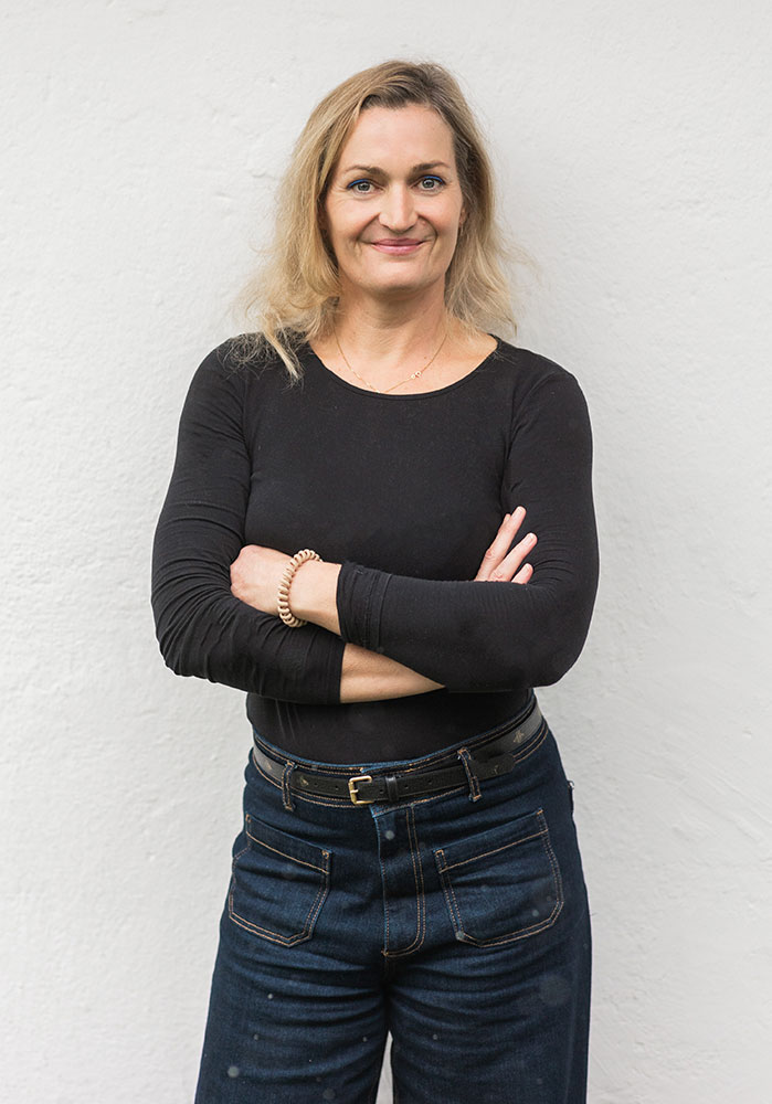 Daniela Burns Daniela Burns steht vor einer hellen wand, die amre lässig verschränkt und lächelt kompetent in die Kamera.